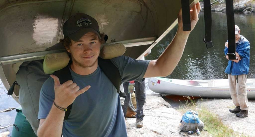 A person balances a canoe on their shoulder and give the camera a "hang loose" sign. 